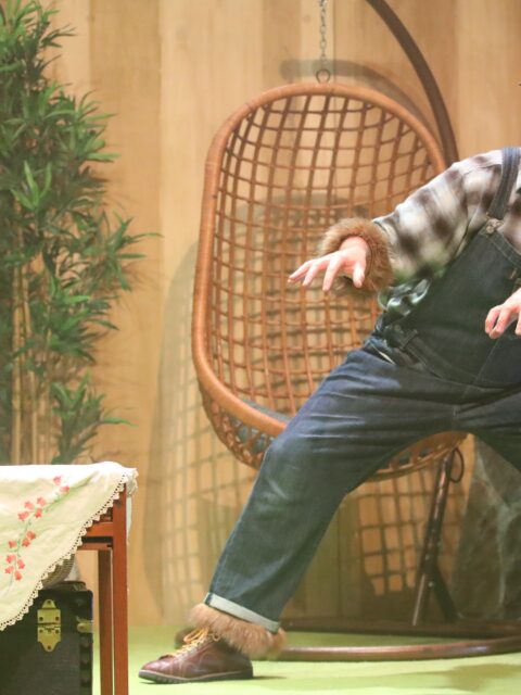 A stage scene from The Three Bears. Grizzly bear, wearing dungarees and a miner's hat with brown fur trim, looks warily at a strange box under a small wooden table. In the background hiding behind a tall plant is Panda bear wearing black and white clothes with a black fur trim.