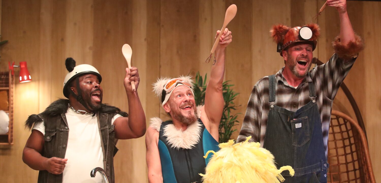 A stage scene from The Three Bears. Three performers are behind a kitchen cabinet. Panda, a bear inspired by a panda, is wearing black and white clothes with a black fur trim. Polar, a bear inspired by a polar bear, is wearing a blue ski suit, ski hat and goggles with white fur trim. Grizzly, a bear inspired by a grizzly bear, wears dungarees and a miner's hat with brown fur trim. Each bear holds up a wooden spoon On the kitchen cabinet are bowls and a large yellow ball of fluff (Goldie).