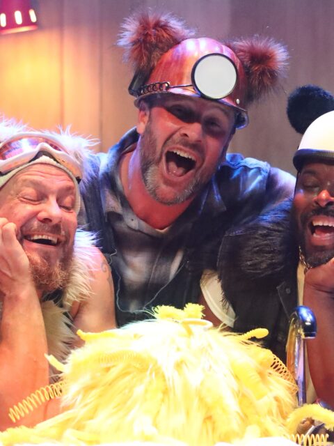 A stage scene from The Three Bears. Three performers are behind a kitchen cabinet. Panda, a bear inspired by a panda, is wearing black and white clothes with a black fur trim. Polar, a bear inspired by a polar bear, is wearing a blue ski suit, ski hat and goggles with white fur trim. Grizzly, a bear inspired by a grizzly bear, wears dungarees and a miner's hat with brown fur trim. They all look lovingly at ‘Goldie’ a mass of bright yellow feathers who is sat infant of them.