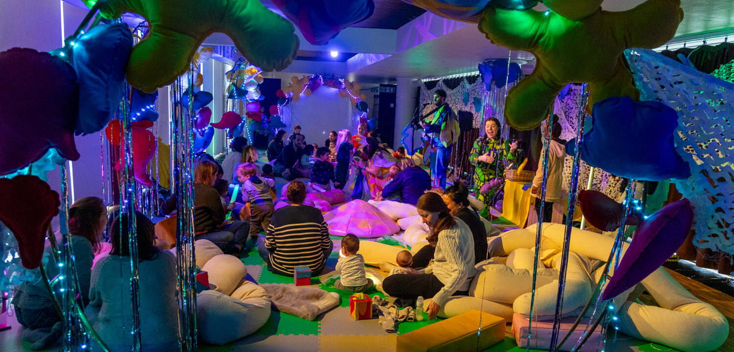 A wide-angle view of a vibrant, immersive sensory room filled with families and children. Large, colorful fabric shapes hang from the ceiling, draped with twinkling blue fairy lights. Parents and toddlers sit on a green and grey foam-tiled floor among oversized soft bolsters. In the background, two performers—one with a guitar—engage with the group under glowing green and purple lights.