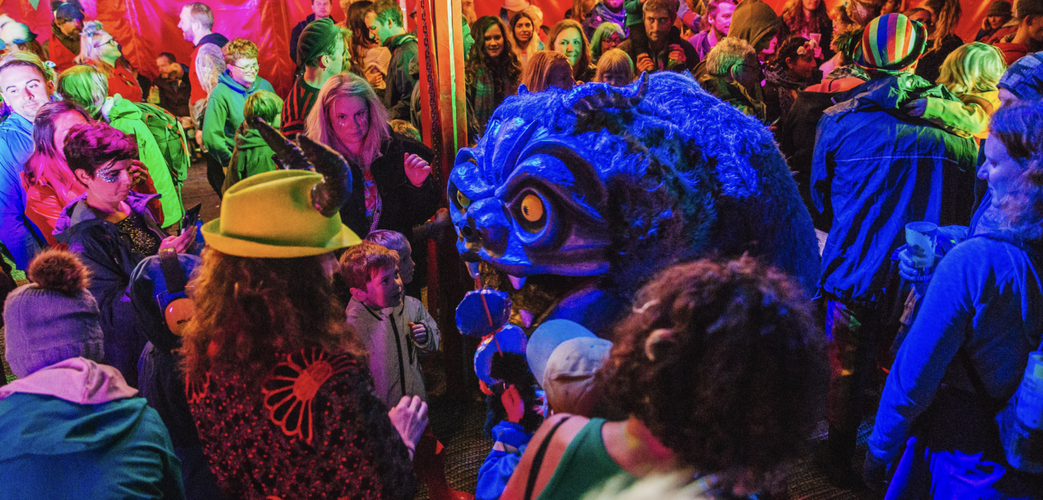 Inside a red circus tent with bunting, a large, shaggy blue monster puppet with glowing yellow eyes stands among a vibrant crowd of children and adults. The scene is filled with colorful stage lighting and people in festive clothing.