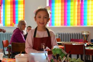 Picture of child smiling directly at the camera, she is wearing a pink jumper and a red apron. In the foreground are paint brushes, egg boxes and other arts and crafts supplies. In the background are rainbow coloured window blinds.