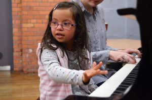 A girl approximately 6 years old is sitting at a piano. She is wearing a grey polkadot jumper and a light pink stripey gilet. Next to her is an adult female wearing a black and white stripey shirt