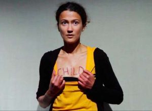 A woman is wearing a black cardigan with a yellow top underneath. She is pulling down the neck of her yellow top to reveal the word 'child' written in pink on her chest.