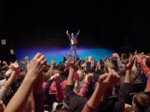Picture of Comedy Club 4 Kids audience - a man stands on stage with his arms raised in front of an audience of children who all have their arms raised in the air.