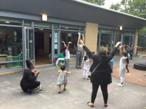 Parents and children dancing together outside. They are all holding their hands up in front of the air and are standing in front of a school building in a playground