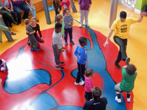 A man wearing a yellow tshirt with 'Capoeira Angola' written on it is kicking his leg in the air. A group of children are watching him. The photo is taken from above and they are standing on the red, blue and yellow abstract patterned floor in Z-arts Atrium space.