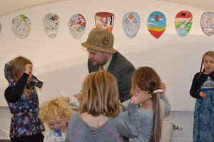 A man wearing a tweed checked pattern bowler hat is holding a book, there are 5 young girls standing around him and some colourful pictures on the white wall behind them