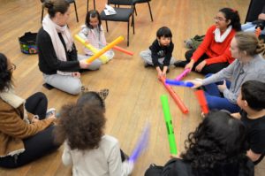A group of children and adults are sat cross-legged on the floor in a circle, they are all holding different brightly-coloured plastic tubes.