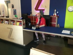 Paul, a member of box office staff is wearing a red Z-arts t-shirt and sitting behind the blue box office desk. He is on the telephone and using a computer.