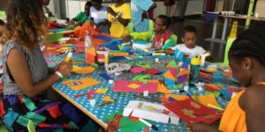 Photo of children and facilitator doing craft activities. There is lots of colourful card on a table covered with a blue and white polkadot tablecloth.