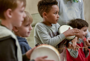 Four children at a music workshop, they are looking off to the right-hand side, one is carrying a tambourine and one is carrying a bongo drum