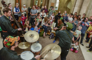A man playing a clarinet, a man playing on a drum kit and a man playing a double bass perform in front of a group of children and families