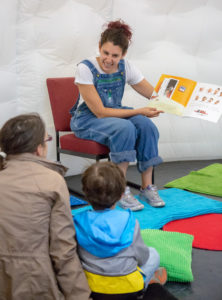 A woman with brown hair wearing denim dungarees and a white t-shirt is sitting reading a book to a group of children.