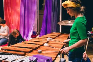 Plink and Boo production picture. A woman in a green top and yellow headband is playing a very large xylophone