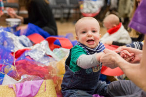 Photo of smiling baby holding an adult's hands. He is wearing a green and grey long-sleeved tshirt and blue denim dungarees. He is sitting in a pile of mixed texture, colourful fabric
