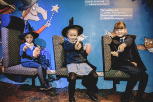 3 young girls sitting in a row, two are wearing witches hats and all three are waving a wand at the camera. In the background is a painting of the witch from 'Room on the Broom'