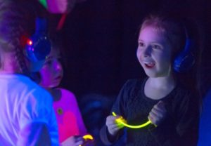 Three children wearing headphones and holding glowsticks. Their clothes are glowing as they are standing under UV light. You can see one child's face clearly, she is blonde and has a happy and surprised expression on her face.