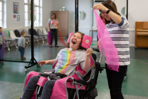 Image of woman who is a wheelchair user laughing with a woman standing next to her with a piece of fabric