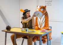 A woman wearing a hat with an orange feather in it is sitting down at a yellow dinner table, she is sitting next to a statue of The Tiger from The Tiger Who Came to Tea and she is holding a plate up to the tiger'sface.