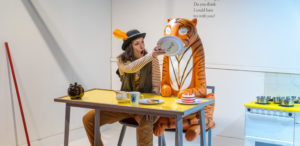A woman wearing a hat with an orange feather in it is sitting down at a yellow dinner table, she is sitting next to a statue of The Tiger from The Tiger Who Came to Tea and she is holding a plate up to the tiger'sface.