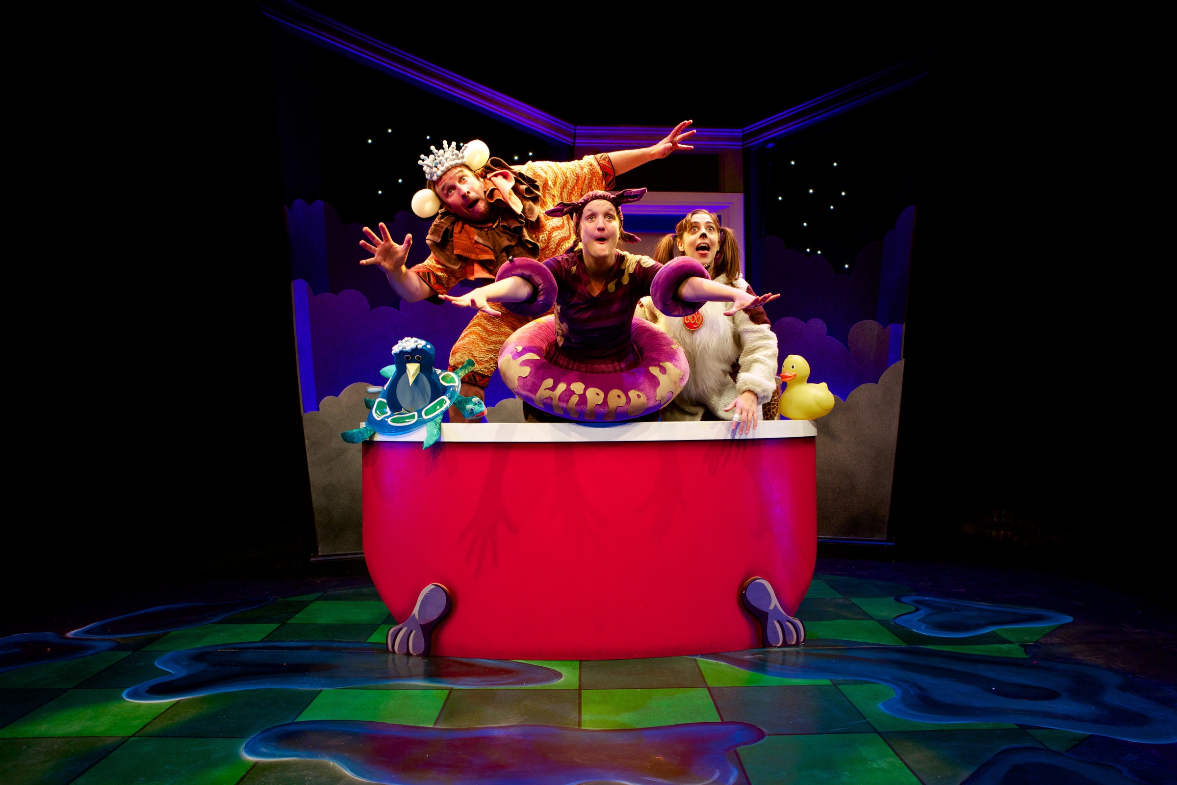 Promo image for Big Red Bath. Three people in costume are standing in a red bath. There is a woman dressed as a dog, a man dressed as a lion and a woman dressed as a purple creature. Two of them are waving and they have looks of amazement on their faces.