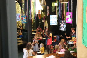 One Manchester Family Jam promo image. A woman is standing singing in front of an audience of young families. She is wearing a colourful dress and there are multicoloured streamers floating down from the ceiling above her head.