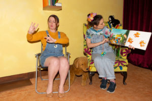 BSL signed performances lead image - Photo from a signed performance of A World Inside a Book. Two women are sat down, one is wearing denim dungarees and holding a book open, she appears to be reading it aloud. The other woman is wearing a denim pinafore dress and is signing in BSL